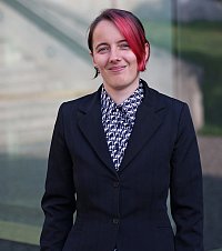Sabine Wller (Photo by Friederike Stecklum)
[Photo description: The photo shows Sabine Wller centered in front of the glass facade of the Audimax. She wears a black and white patterned shirt and a black blazer, has brown hair and a red streak. She looks directly into the camera and smiles.]