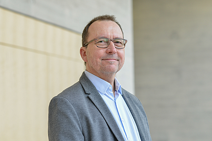 Photo: Götz-Olaf Wolff (Photographer: Markus Scholz) [Image Discription: Götz-
Olaf Wolff stands in the Audimax and looks smiling into the camera. He wears 
glasses, a gray suit and a light blue shirt and has short brown hair].